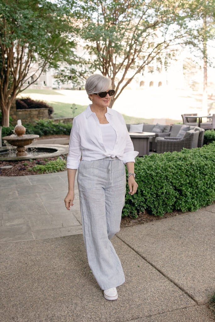 Woman with short silver hair wearing a white button-front shirt tied at the waist over a white tank, paired with light gray linen wide-leg pants, white sneakers, and oversized sunglasses while walking outdoors near a fountain and landscaped patio.