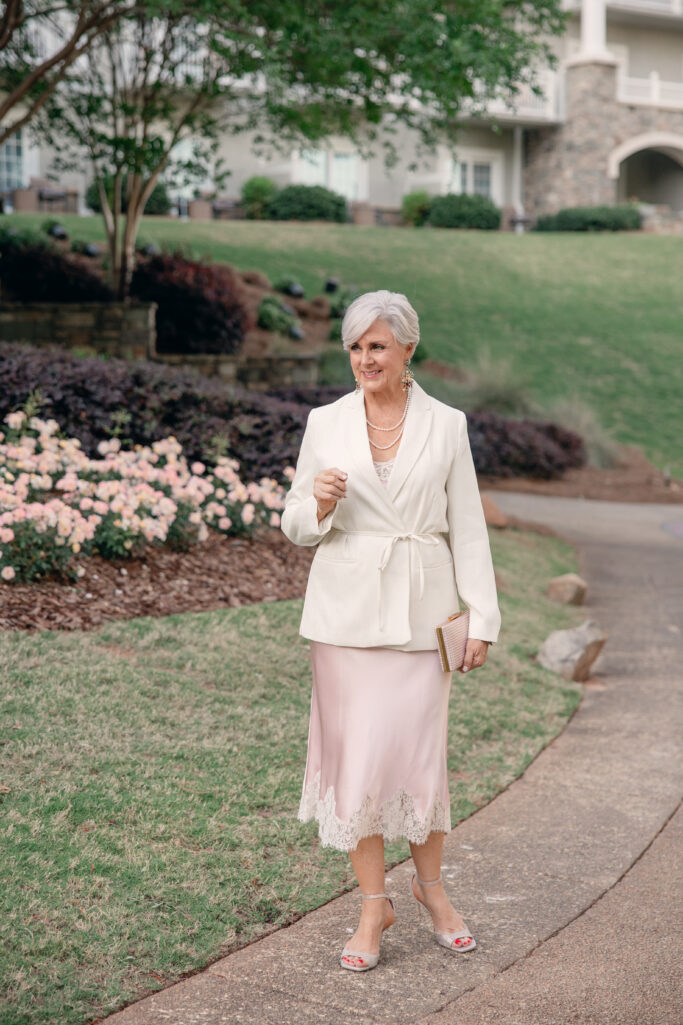 Woman over 50 wearing a blush lace skirt and ivory belted blazer for a wedding guest outfit.