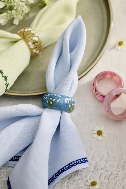 Spring-inspired table setting featuring pastel linen napkins, hand-painted floral napkin rings, a woven rattan charger plate, and fresh white flowers arranged on a neutral tablecloth.