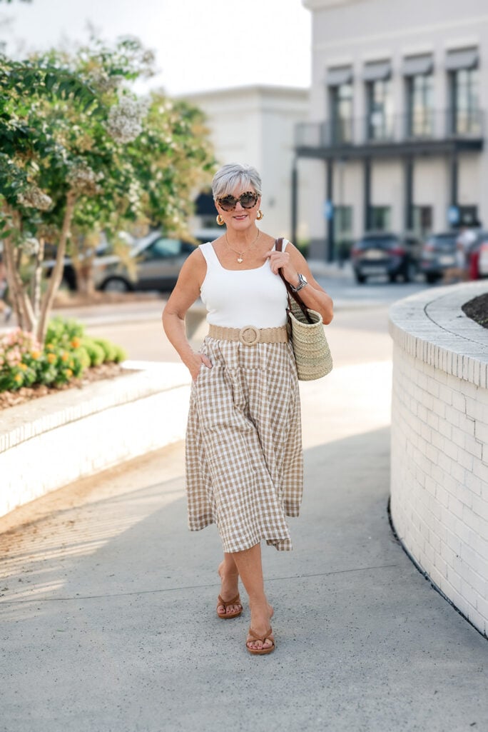 Woman over 50 styling a gingham midi dress, woven belt, and straw market tote