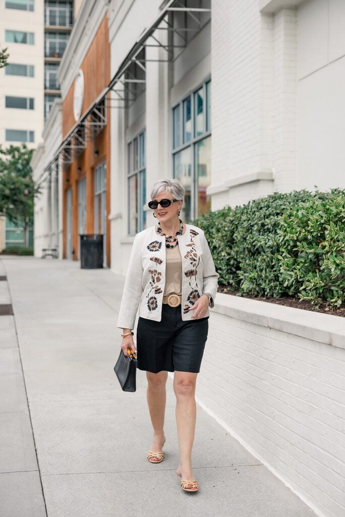 Beth of Style at a Certain Age wearing an embroidered linen jacket with a tan tank and black Bermuda shorts, carrying a black top-handle bag — one of the best summer handbags for women over 50