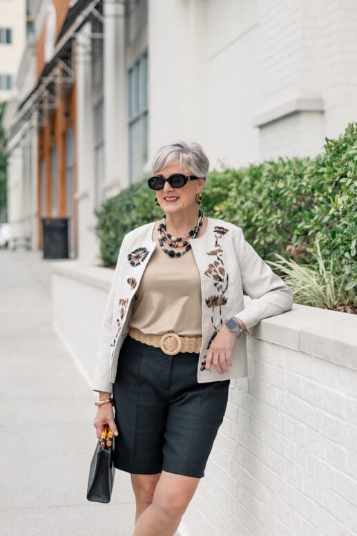 Beth of Style at a Certain Age smiling and leaning against a low wall, wearing black oval sunglasses, a layered black and ivory beaded necklace, gold drop earrings, an ivory floral-embroidered jacket, a beige satin tank, a woven straw belt, black bermuda shorts, a smartwatch, and carrying a black leather bag with wooden handles, on a city sidewalk