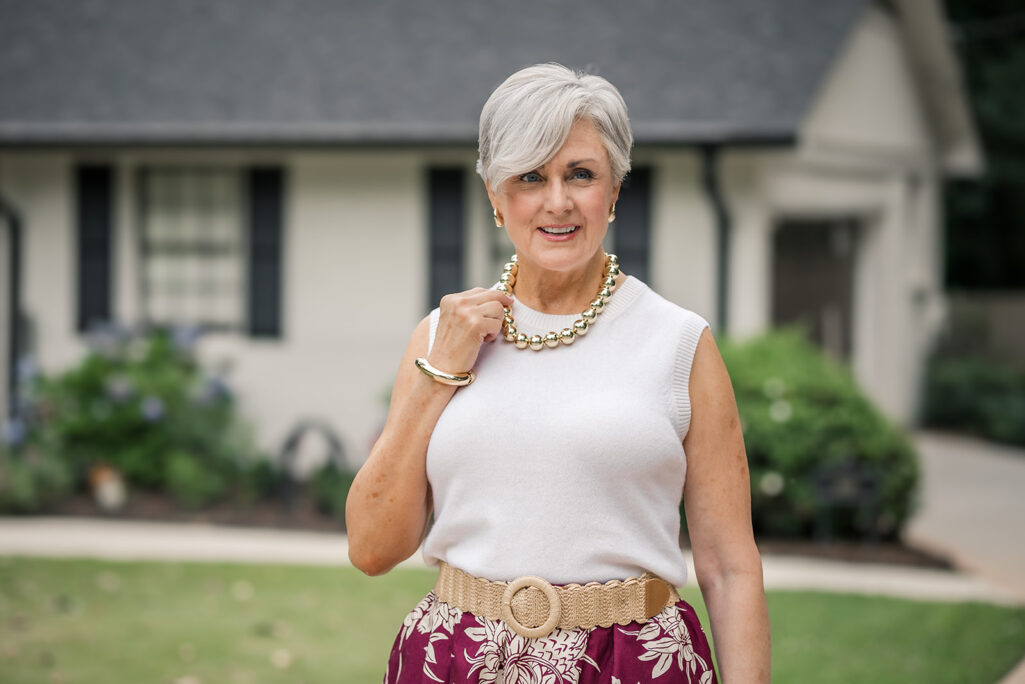 Beth of Style at a Certain Age facing forward and smiling, wearing a chunky gold ball bead necklace, small gold stud earrings, and a gold dome cuff bangle, styled with a white sleeveless sweater tank, burgundy floral midi skirt, and a woven raffia belt with a round wooden buckle