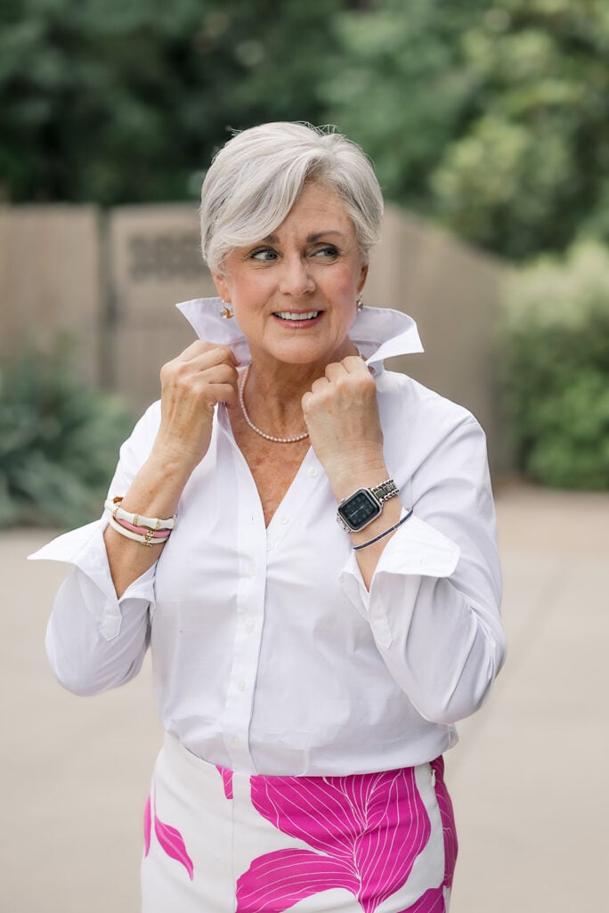 Beth of Style at a Certain Age in a white button-down shirt and pink floral shorts, popping the collar with both hands, wearing a colorful mixed bracelet stack with pink leather or cord, white enamel bangle, and gold chain on her right wrist, Apple Watch with silver link band on her left