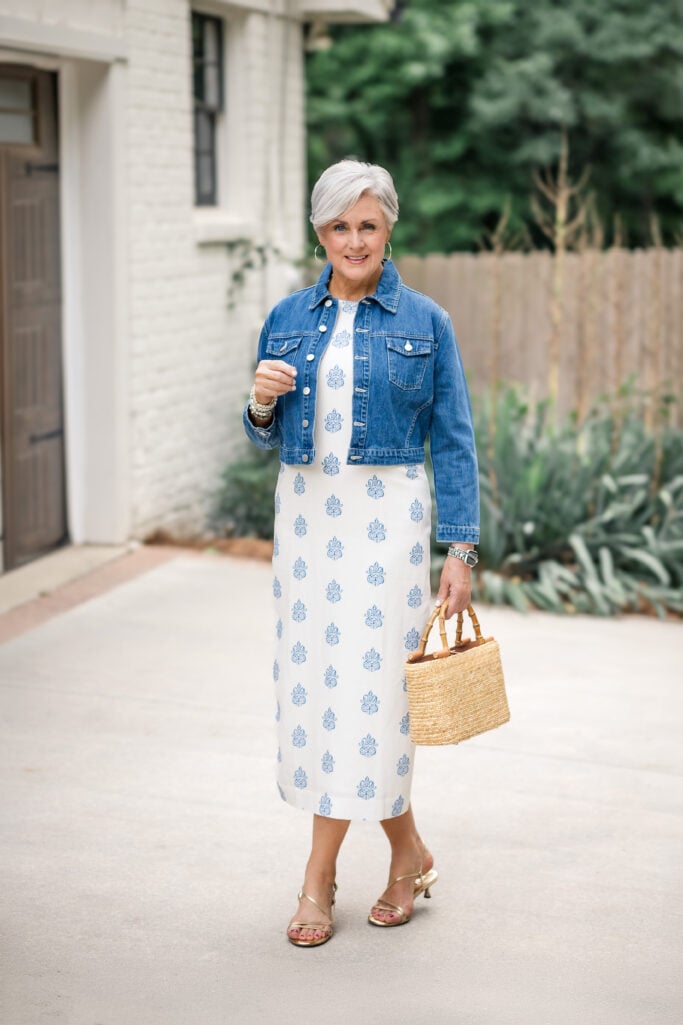 Beth wearing a white midi dress with a blue block print, cropped denim jacket, gold strappy heels, and a straw bag with bamboo handles