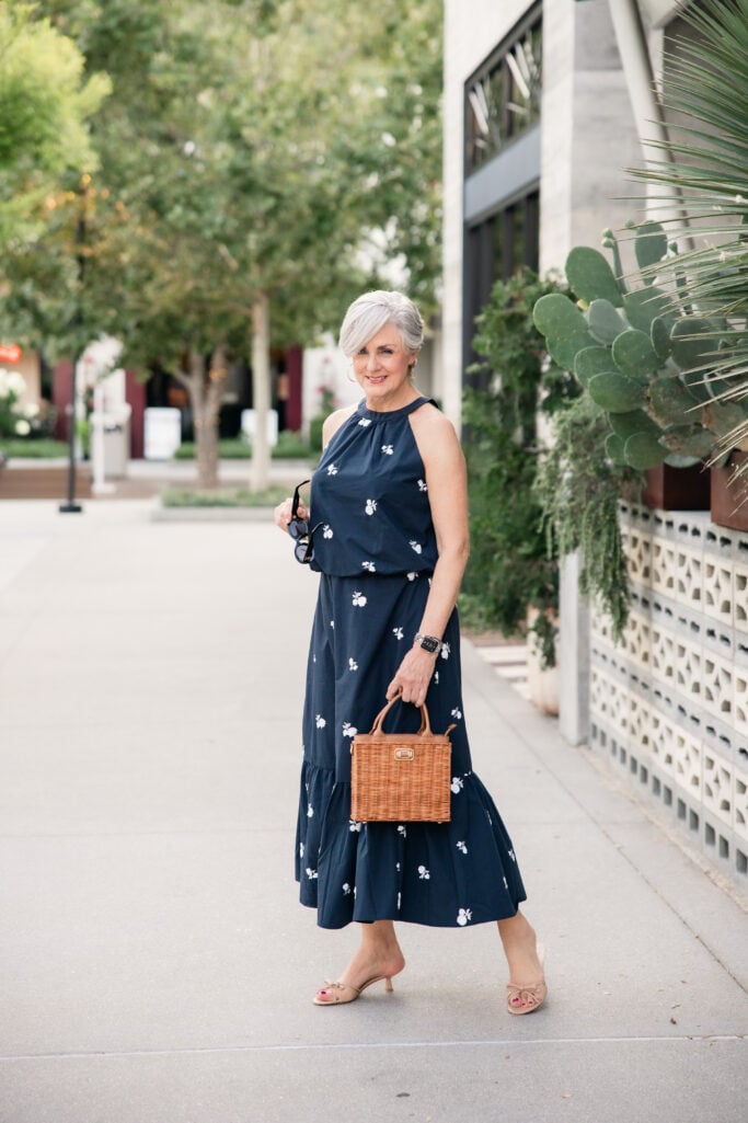 Navy halter maxi dress with white embroidered florals, wicker handbag, and nude kitten heels