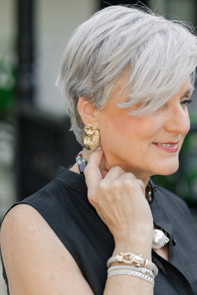 Close-up of Beth of Style at a Certain Age wearing a large sculptural gold oyster-shaped clip earring, with layered bracelets on her wrist, styled with a black sleeveless linen top
