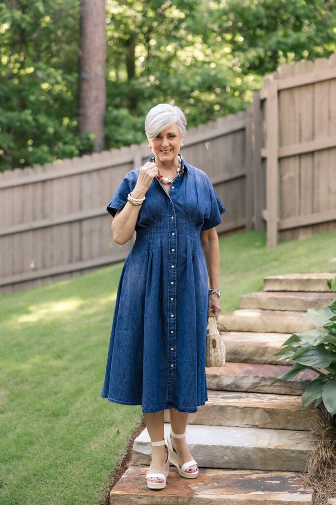 Beth of Style at a Certain Age in a dark wash denim midi shirtdress standing on garden steps, wearing a stack of gold bangles on her right wrist, pearl earrings, and a layered necklace, holding a small wicker basket bag