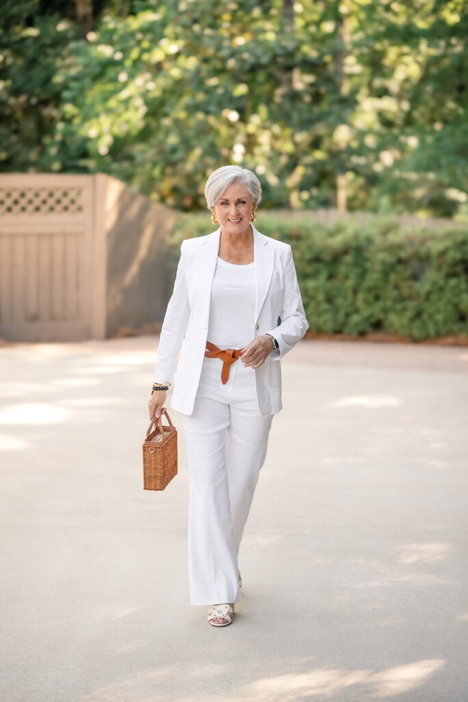 Beth of Style at a Certain Age in an all-white single-button blazer and wide-leg white trousers, styled with a white tee, brown leather tie belt, small structured wicker box bag, white strappy sandals, and gold hoop earrings.