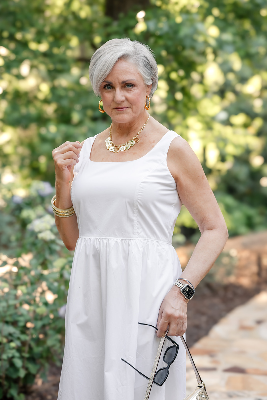 Beth of Style at a Certain Age in a white square-neck midi dress holding sunglasses, wearing a gold coin disc chain necklace, large gold resin hoop earrings, and stacked gold bangles