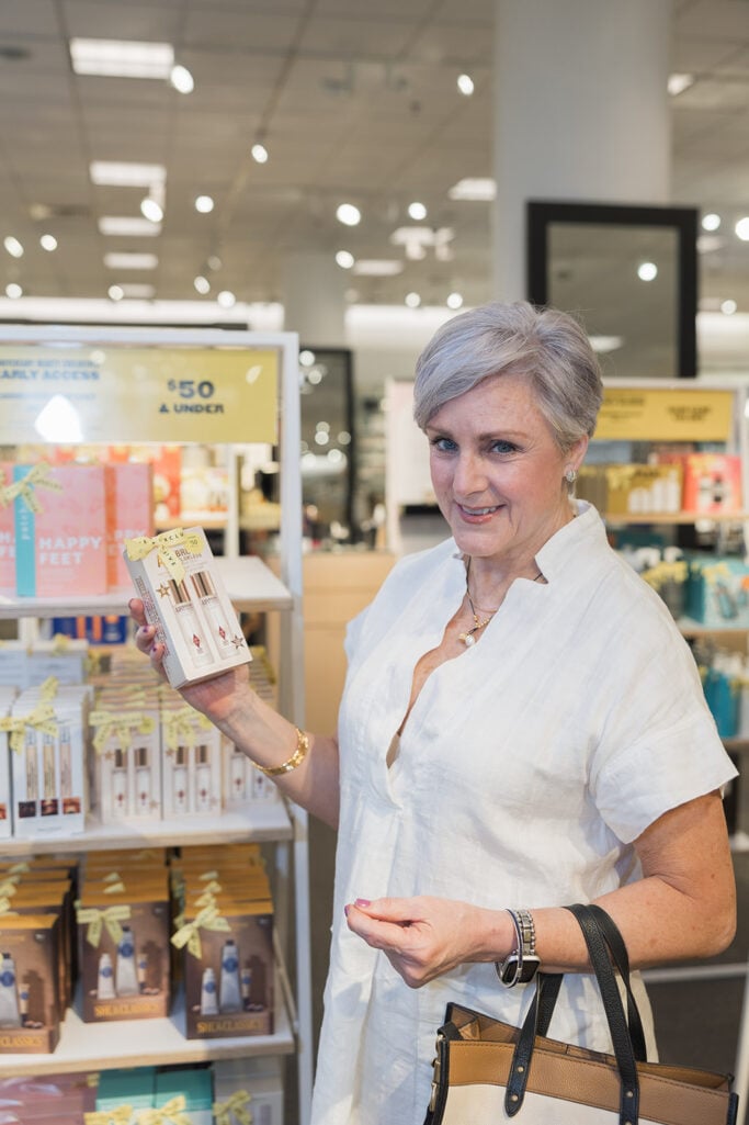 Beth of Style at a Certain Age smiling and holding a boxed beauty set at the Nordstrom Anniversary Sale beauty counter, wearing a white linen dress and gold jewelry, with a $50 and under display shelf in the background.