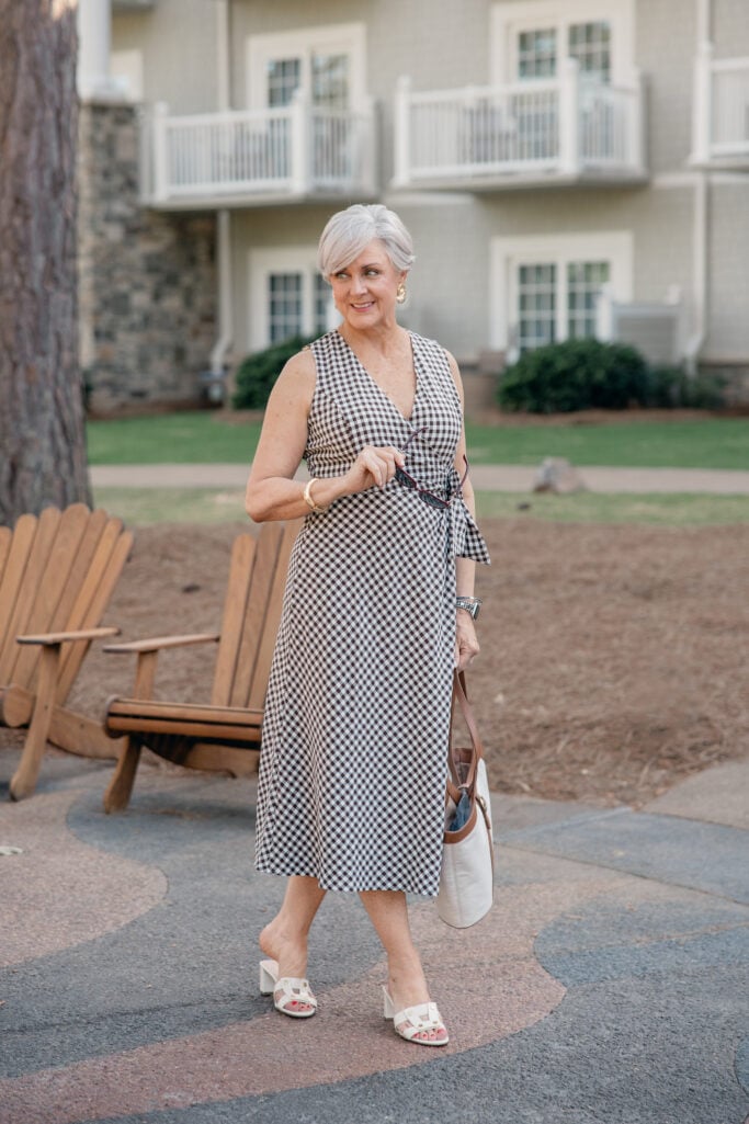 Beth wearing a brown gingham sleeveless wrap midi dress with white heeled slides at a resort