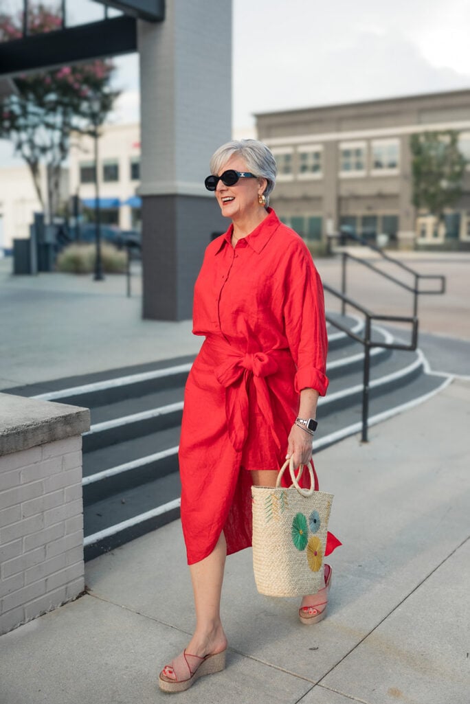 Woman over 50 in a red linen Apiece Apart shirtdress with a straw tote and espadrille wedges — an easy Friday outfit for a reunion weekend.