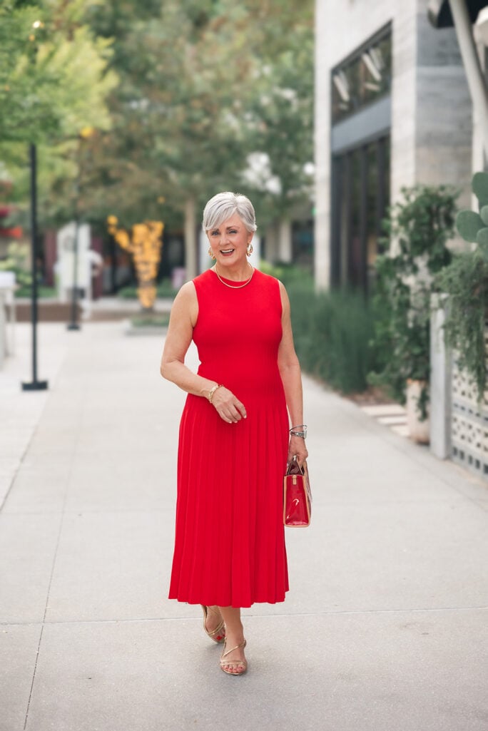 Woman over 50 in a red pleated midi dress with gold sandals and a gold bag — a cocktail outfit for a high school reunion.