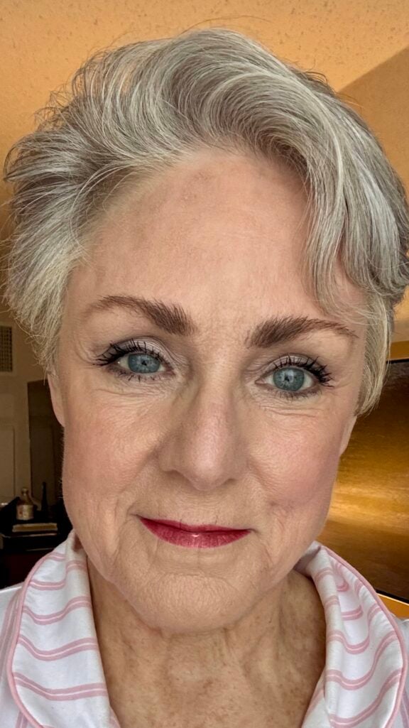 Close-up of a woman over 50 with silver hair, soft glowy skin, a neutral eye, and a rosy-mauve lip — natural daytime makeup for a reunion.