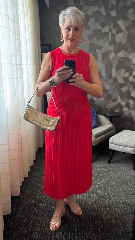 Silver-haired woman in a red pleated midi dress and gold strappy sandals, holding a gold metallic bag on a city sidewalk — reunion cocktail outfit for women over 50.