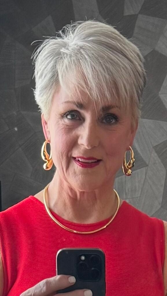 Closeup of Woman over 50 with short silver hair wearing a sleeveless red pleated midi dress, gold jewelry, and gold heels — what to wear to a high school reunion cocktail party.