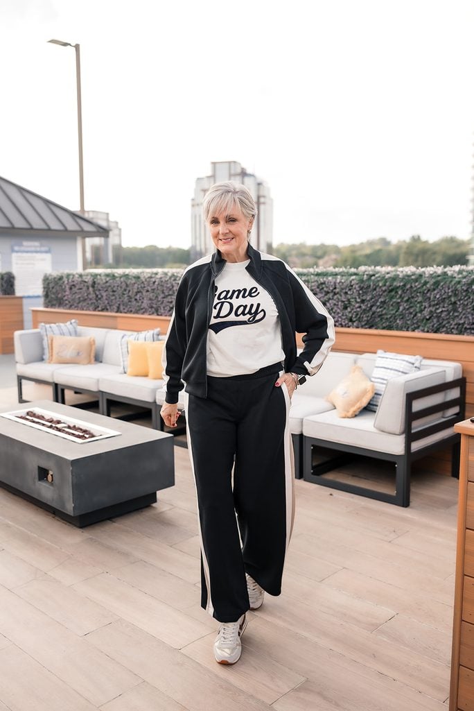 Beth Djalali in a black track jacket with cream side stripes over a Game Day tee with matching wide-leg track pants and metallic sneakers