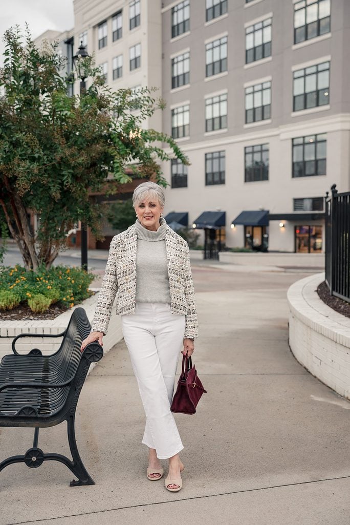 Beth Djalali in one of her fall 2026 outfits for women over 50 — a cream tweed jacket worn open over a gray turtleneck with white straight-leg jeans and a burgundy suede bag