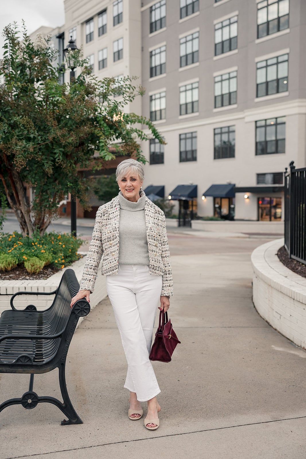 Beth Djalali in one of her fall 2026 outfits for women over 50 — a cream tweed jacket worn open over a gray turtleneck with white straight-leg jeans and a burgundy suede bag