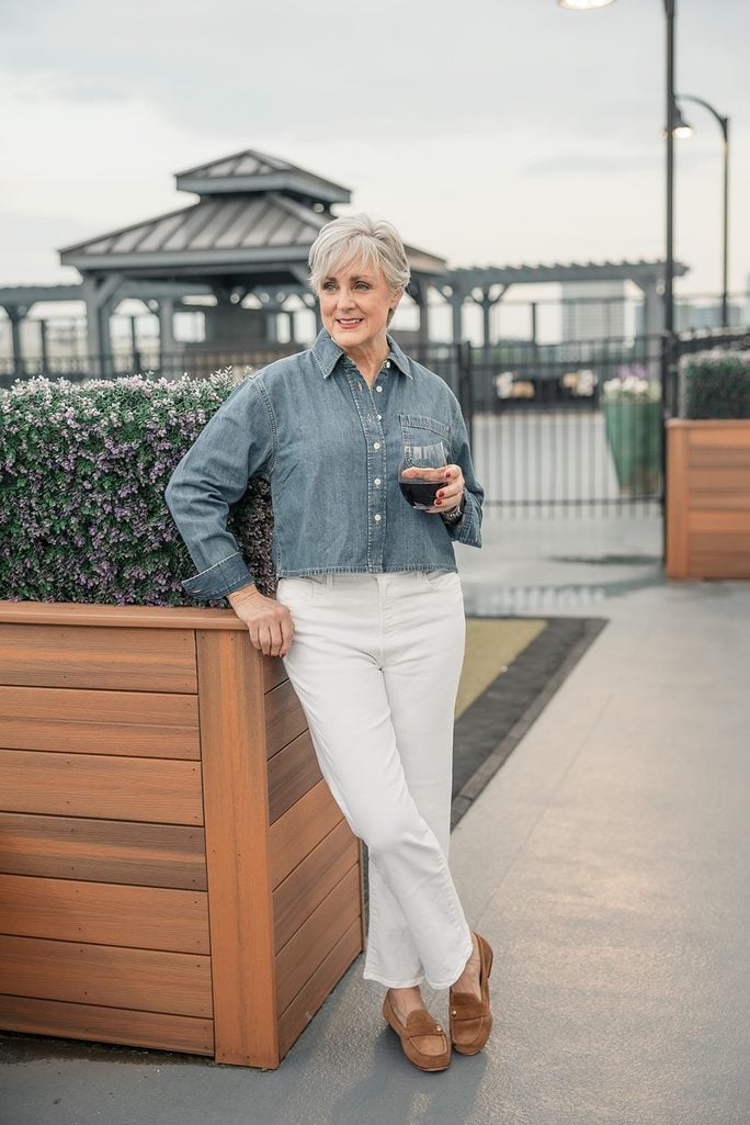 Beth Djalali in a boxy cropped denim shirt, white jeans, and cognac suede loafers on a rooftop terrace