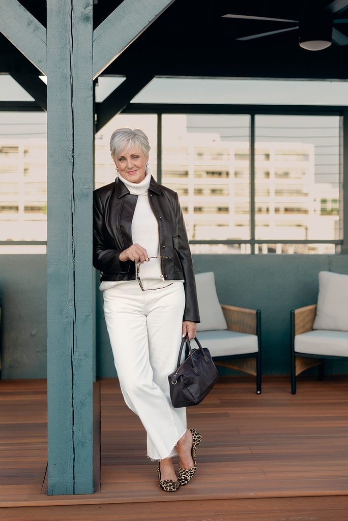 Beth leaning against a teal pergola post in a black faux leather jacket over a white turtleneck, white wide-leg jeans and leopard flats, holding a black leather satchel