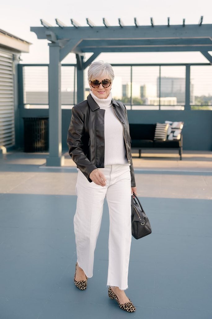Beth walking across a rooftop terrace in a cropped black faux leather jacket, cream turtleneck, frayed-hem white jeans and leopard buckle flats, tortoiseshell sunglasses on