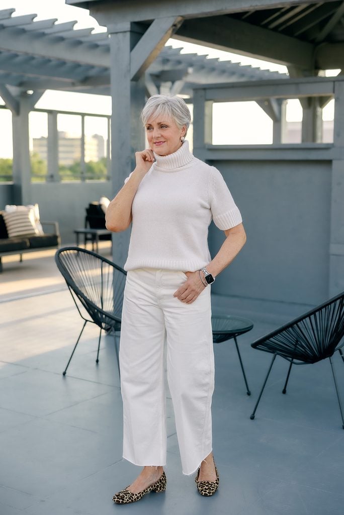 Beth in a white short-sleeve turtleneck sweater and white barrel jeans with leopard buckle flats on a rooftop deck, black satchel in hand