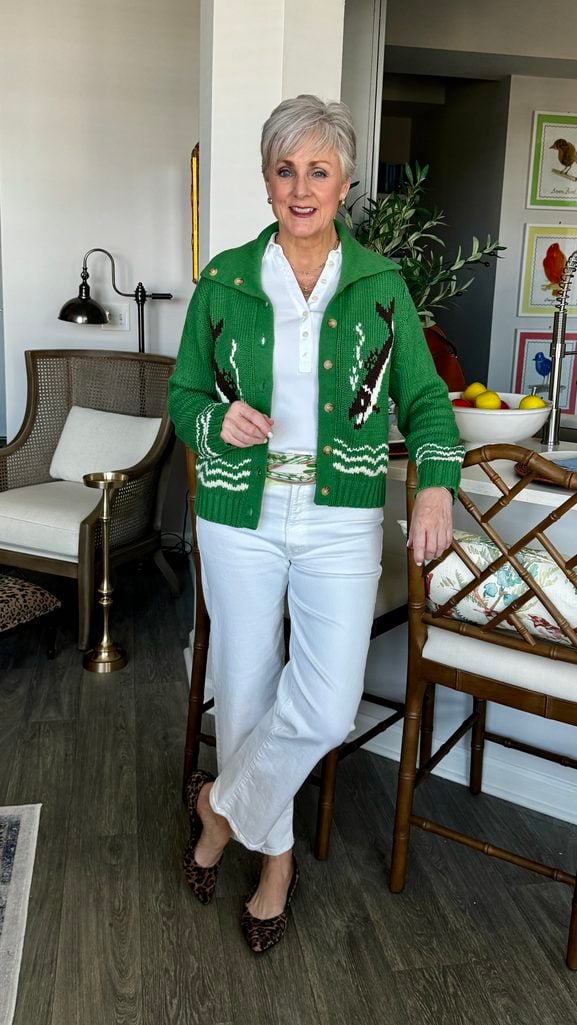 Beth in a green fish-motif cardigan over a white polo, white wide-leg jeans, and leopard pointed flats.