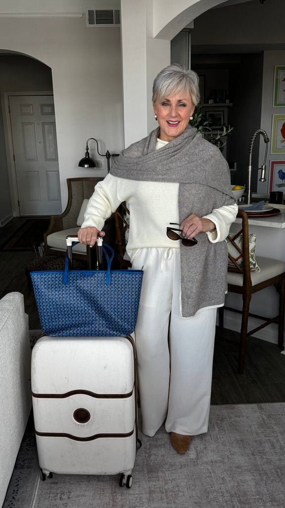 Beth Djalali in a gray cashmere wrap over an ivory sweater and ivory wide-leg pants with a blue tote on a white suitcase