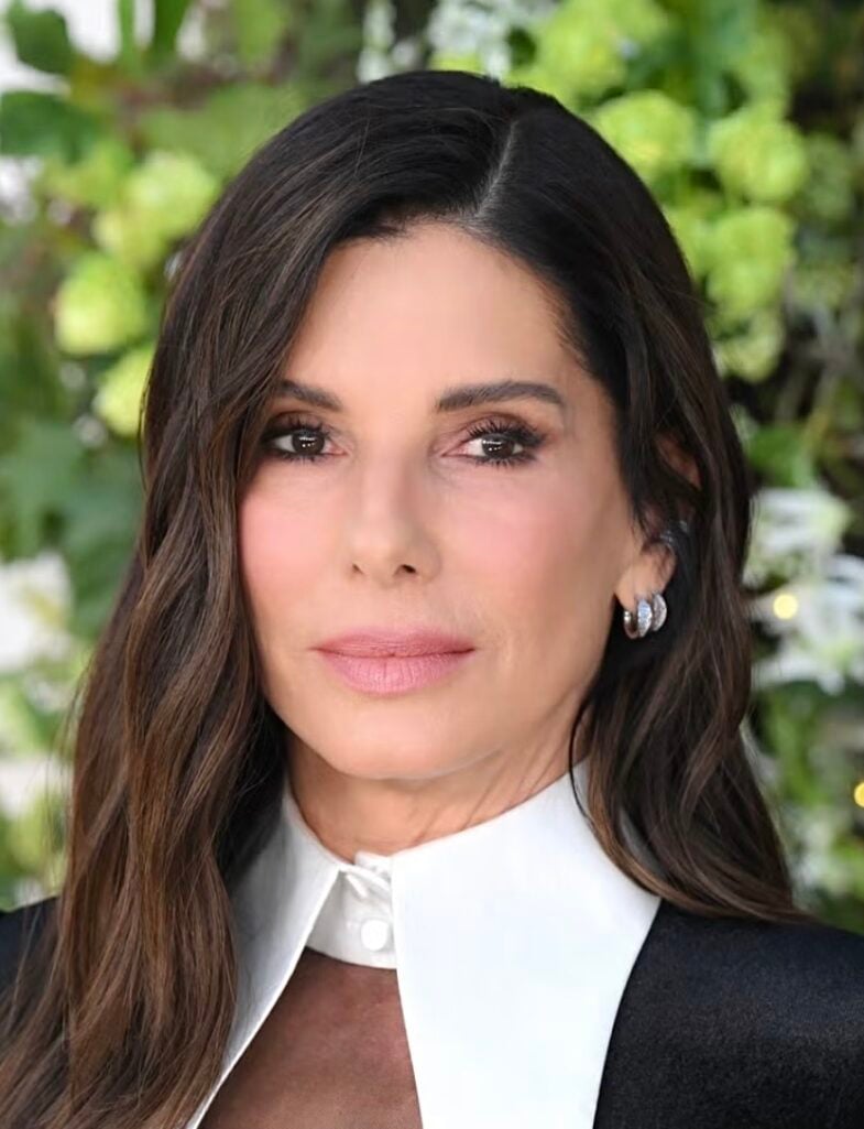 Close-up of Sandra Bullock with dark waves, a white collared top, light smoky brown eyes with black liner, and a satin nude-pink lip
