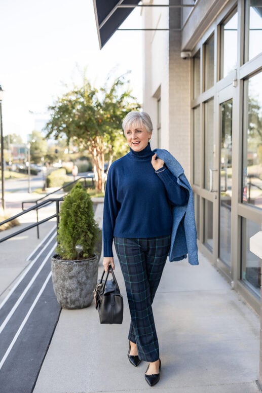 When to start holiday shopping: Beth Djalali in a navy cashmere turtleneck, blackwatch plaid pants and black pointed-toe flats, carrying a black satchel with a blue bouclé jacket over her shoulder.