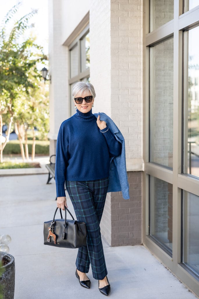 Beth Djalali in a navy cashmere turtleneck, blackwatch plaid pants and black pointed-toe flats, carrying a black satchel with a blue bouclé jacket over her shoulder.