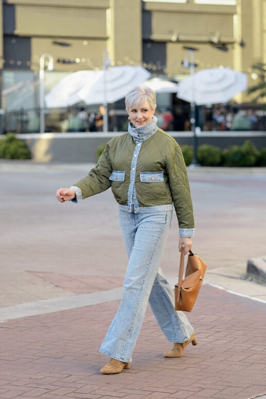Beth Djalali walking in an olive quilted jacket with denim trim, light-wash high rise flare jeans, tan suede boots and a cognac T-Lock handbag