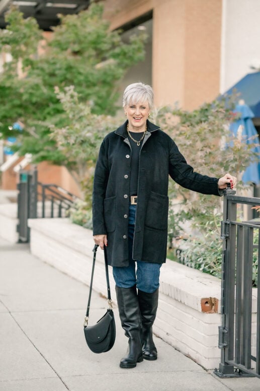 Beth Djalali in a black reversible coat, black crewneck sweater, dark-wash jeans with a gold-buckle belt, and tall black riding boots, holding a black saddle bag on a city sidewalk