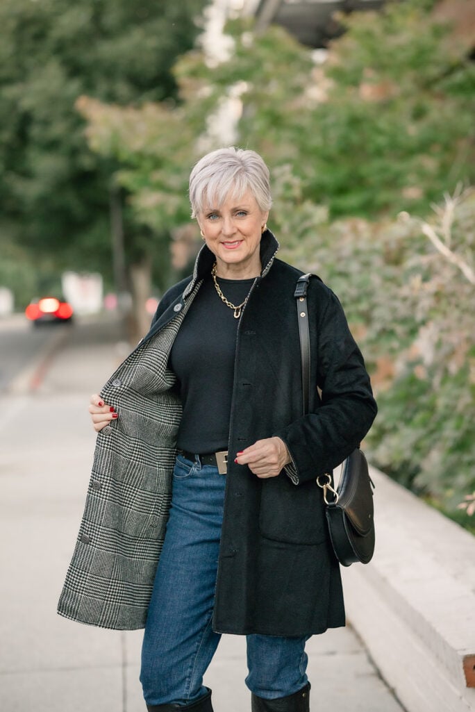 Beth Djalali holding open a black reversible coat to show its black-and-white glen plaid side, worn with a black sweater, gold chain necklace, dark jeans, and a black saddle bag