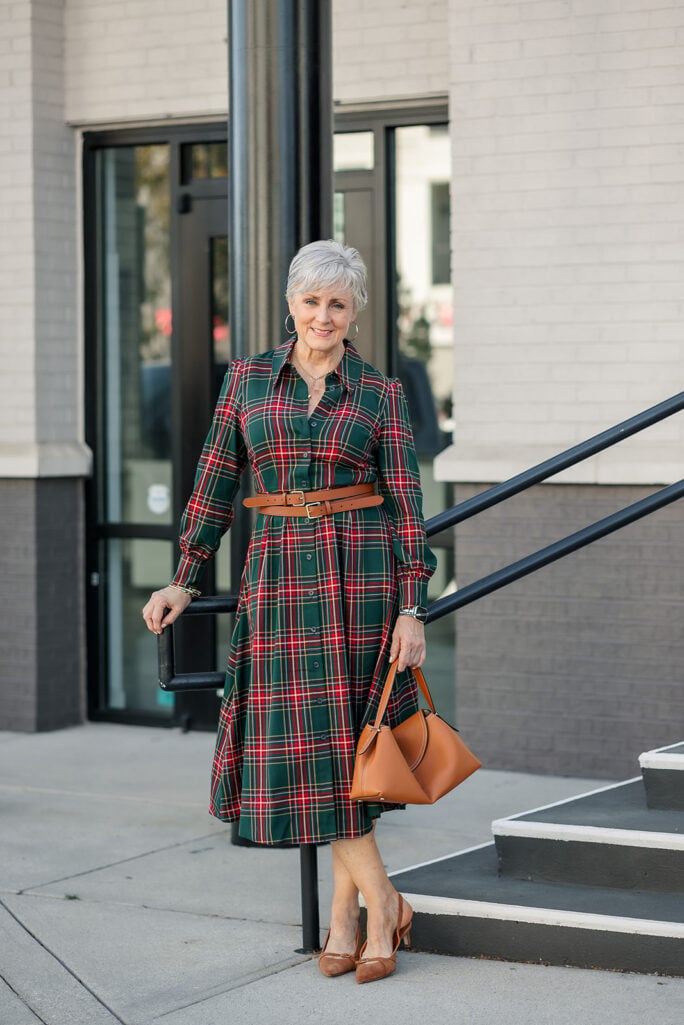Beth Djalali in a tartan shirtdress holding a cognac leather handbag beside a black stair railing