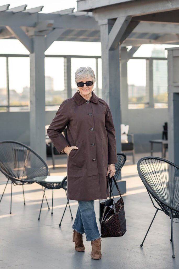 Beth Djalali on a rooftop terrace in a brown mac with a corduroy collar, straight leg jeans, brown suede ankle boots, oval sunglasses, gold hoops and a leopard patent tote