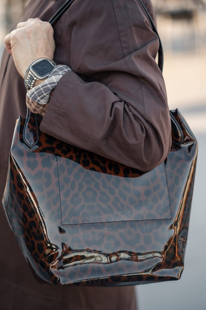 Close-up of a leopard patent tote carried on the shoulder with a brown mac, plaid shirt cuff and silver watch