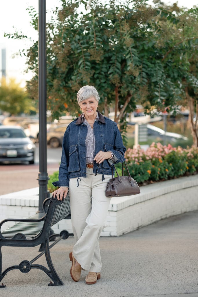 Casual fall outfit for women over 50: Beth Djalali of Style at a Certain Age in a dark denim jacket, gingham shirt, cream chinos and tan suede loafers, leaning on a park bench