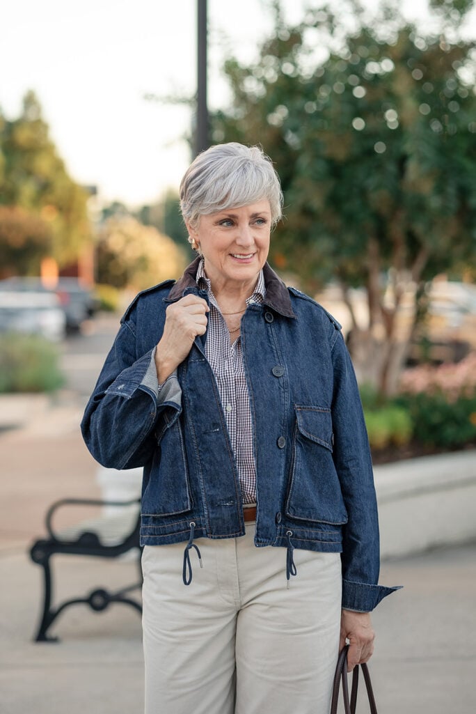 Beth Djalali smiling in a dark denim jacket over a brown gingham shirt with layered gold necklaces and cream chinos
