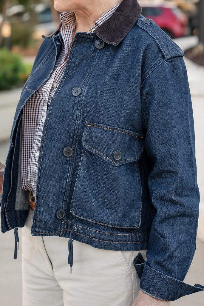 Beth adjusting black oval sunglasses, wearing a dark wash denim jacket with a brown corduroy collar over a navy gingham shirt, carrying a dark brown leather top-handle bag