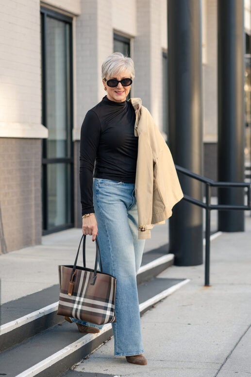 Beth of Style at a Certain Age wearing a cropped trench coat draped over one shoulder, a black funnel neck top, straight leg jeans, and carrying a brown and beige check tote bag