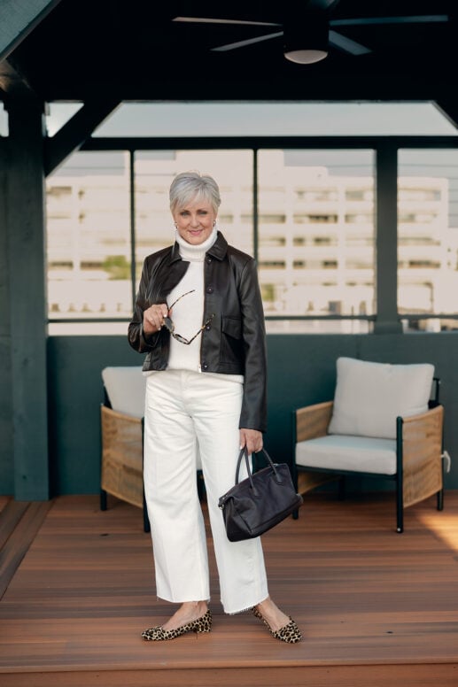 Beth of Style at a Certain Age standing on a covered rooftop deck holding tortoiseshell sunglasses, wearing a dark brown leather jacket, cream turtleneck sweater, ecru wide-leg jeans and leopard flats, carrying a dark leather top-handle bag