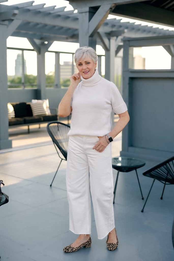 Beth in a cream short-sleeve turtleneck sweater, ecru barrel jeans with a raw hem, and leopard print flats on a rooftop terrace.