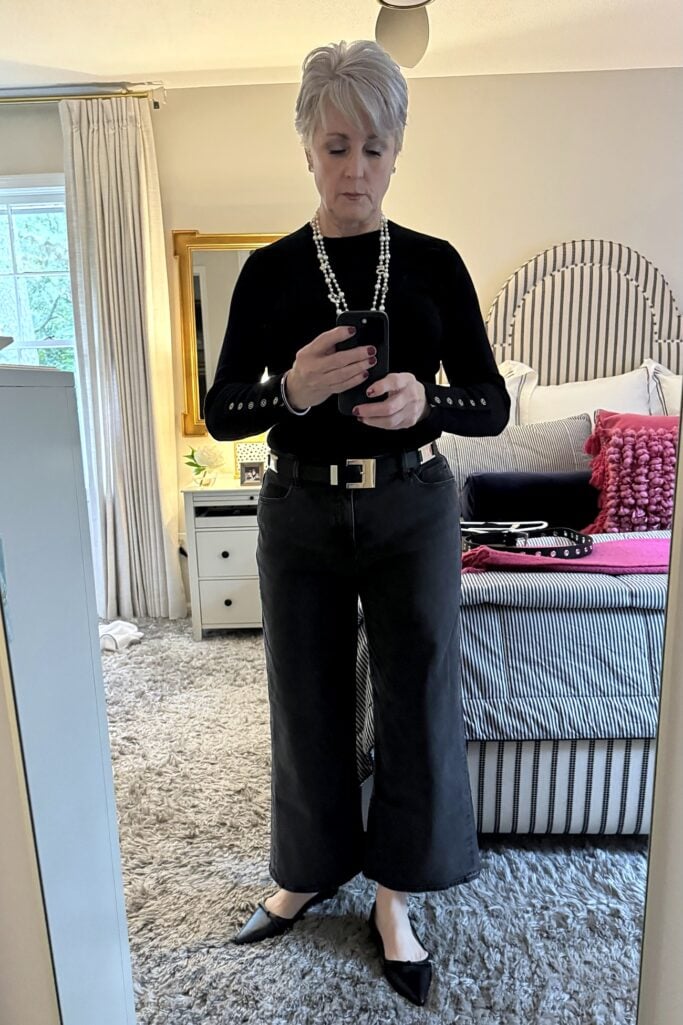 Beth in black sweater, wide black jeans and pearls