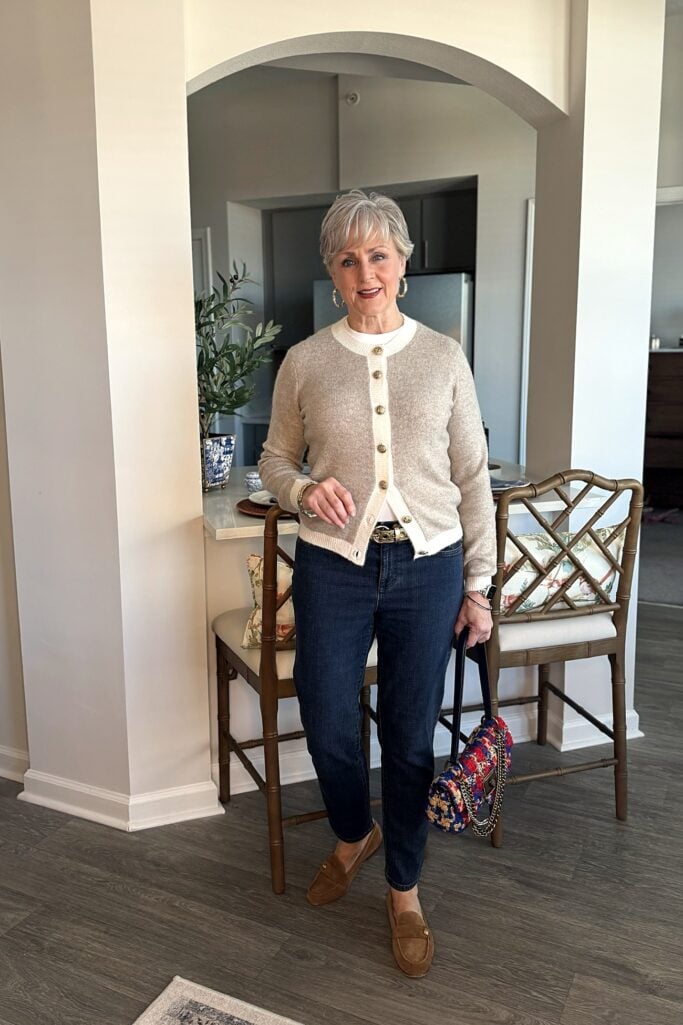 Beth Djalali wearing a cozy chic outfit over 50: an oatmeal cardigan with cream trim and gold buttons, dark-wash jeans, tan suede loafers and a tweed chain bag at home