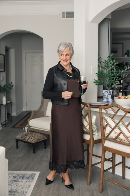 Date night outfit over 50: Beth holding a glass of red wine in a chocolate lace-trim cami and midi skirt, black leather knit jacket, pearls and slingback heels