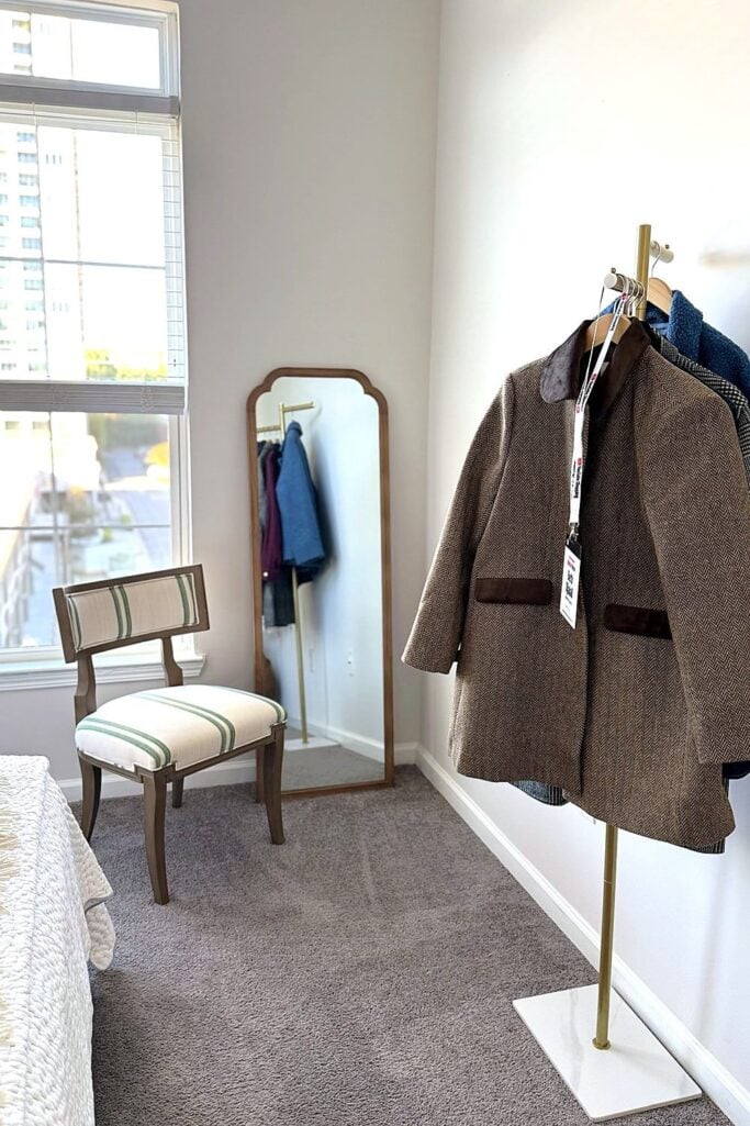 Striped upholstered chair, arched standing mirror, and brass clothes rack in a sunny bedroom corner