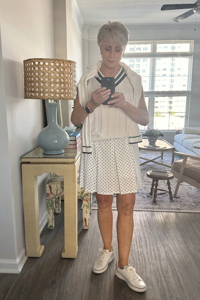 Mirror selfie of Beth in her Atlanta apartment wearing a cream cable-knit tennis vest, a cardigan tied over her shoulders, and a polka dot pleated skort, next to a cane-shade lamp and grasscloth console table